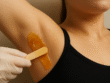Close-up de uma axila feminina sendo depilada com cera de mel aplicada com uma espátula de madeira por uma profissional usando luva de proteção.